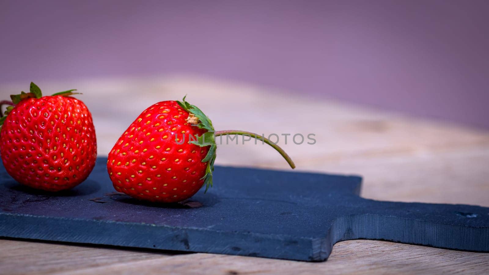 Close up of two strawberries on small black cutting board isolated outdoor on wooden table.