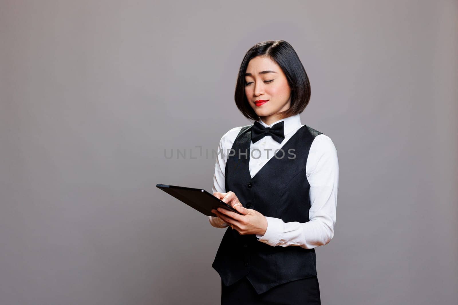 Restaurant administrator young asian woman using digital tablet while posing in studio. Attractive waitress wearing professional uniform and bow tie holding portable device