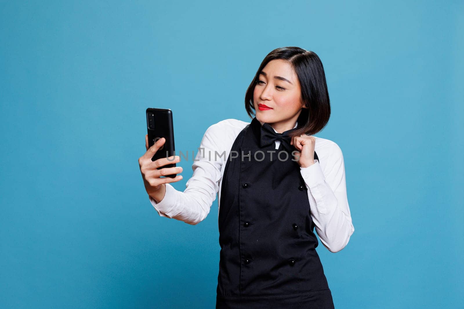 Attractive asian waitress adjusting bow tie and looking at smartphone front camera while taking selfie. Restaurant woman employee in uniform posing making photo on mobile phone