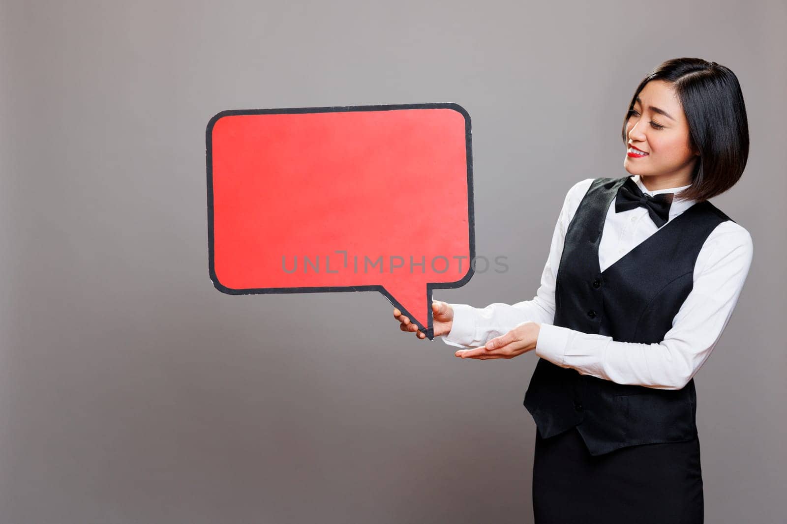 Smiling asian waitress showing red empty message banner mockup for restaurant advertising. Cafe woman employee in uniform holding blank chat bubble with copy space for promotion