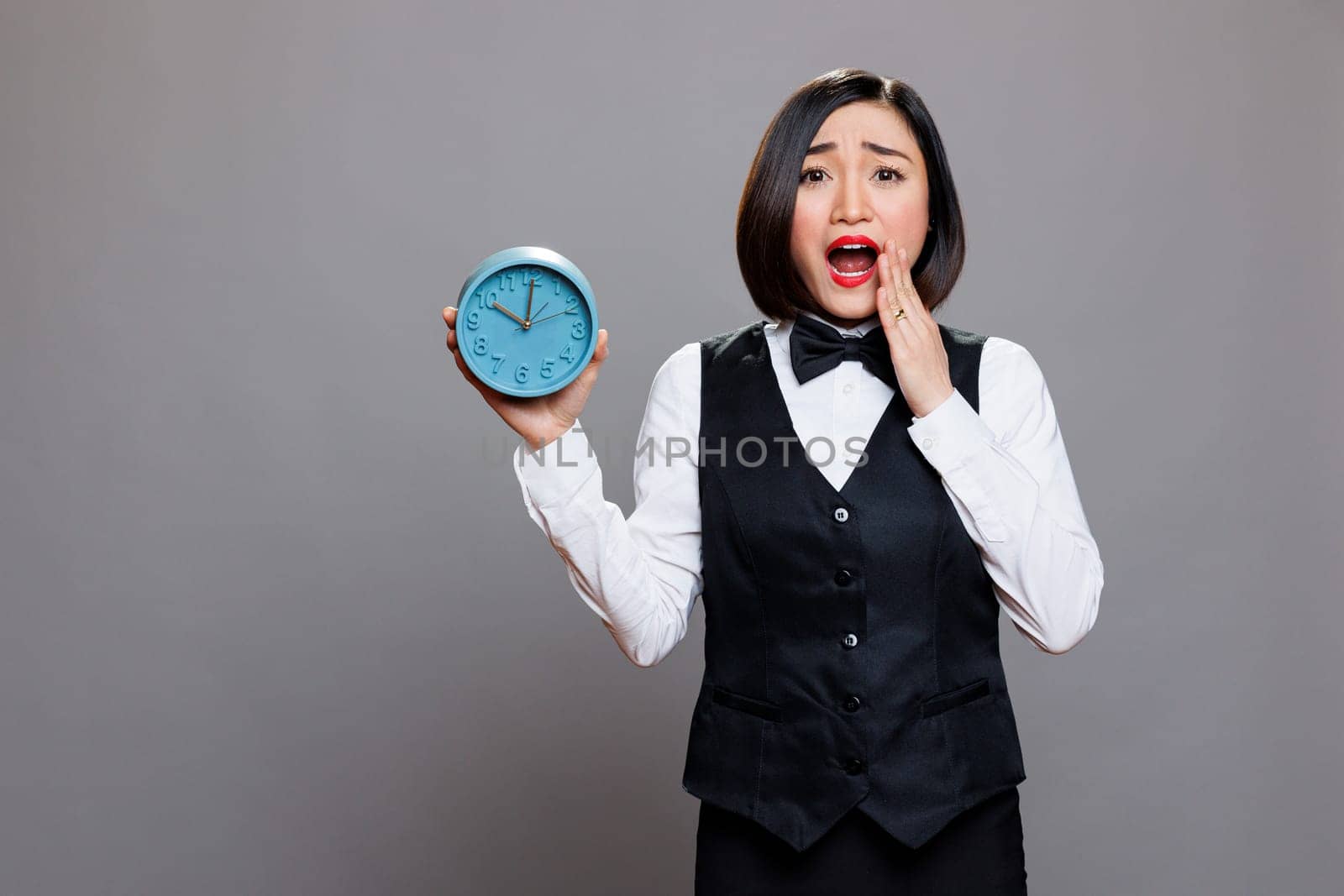 Anxious asian waitress panicking while holding circle dial alarm clock and looking at camera with open mouth. Young terrified woman receptionist showing timer watch portrait