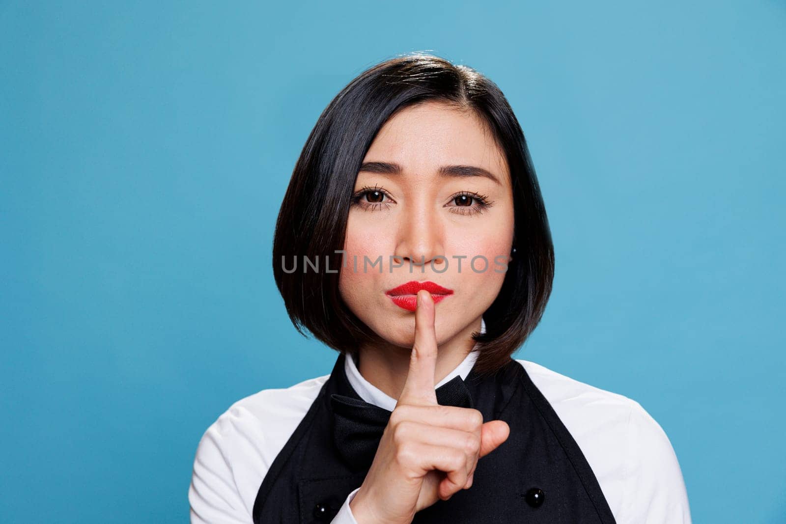 Hotel young asian woman receptionist making silent gesture, asking to keep secret portrait. Restaurant waitress wearing uniform holding forefinger on lips and looking at camera