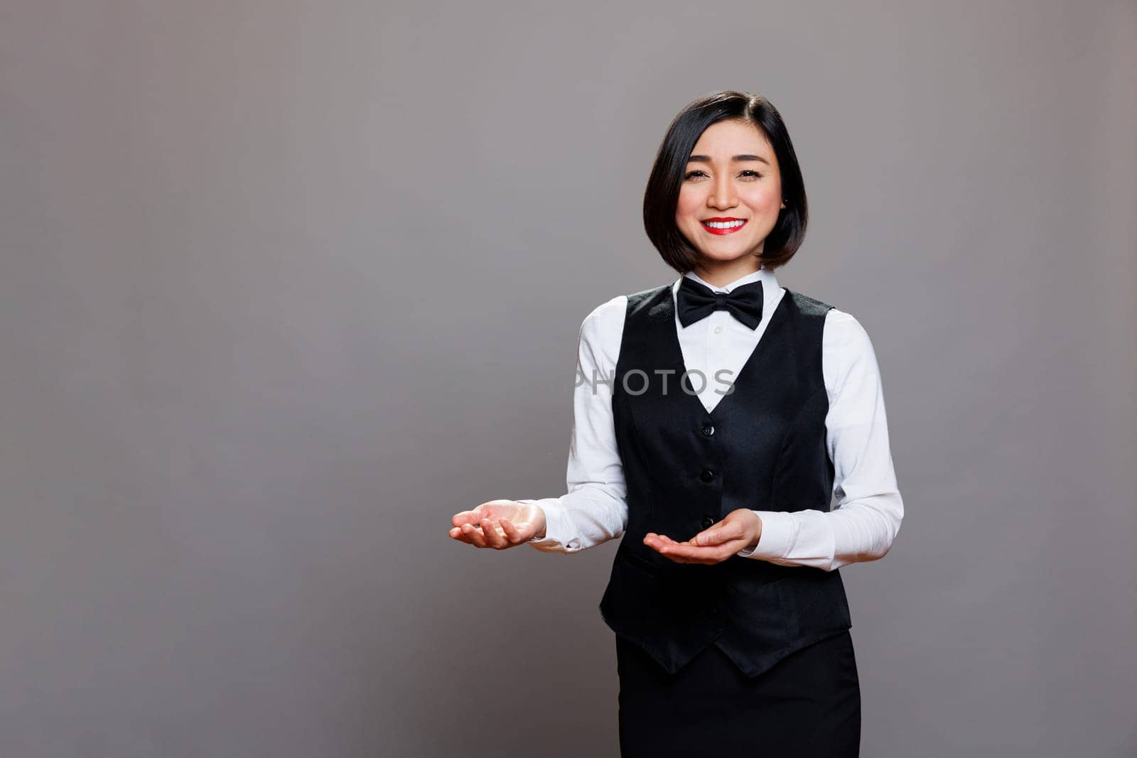 Smiling asian woman restaurant receptionist welcoming guests and showing with hands portrait. Friendly waitress in uniform greeting customers while posing and looking at camera