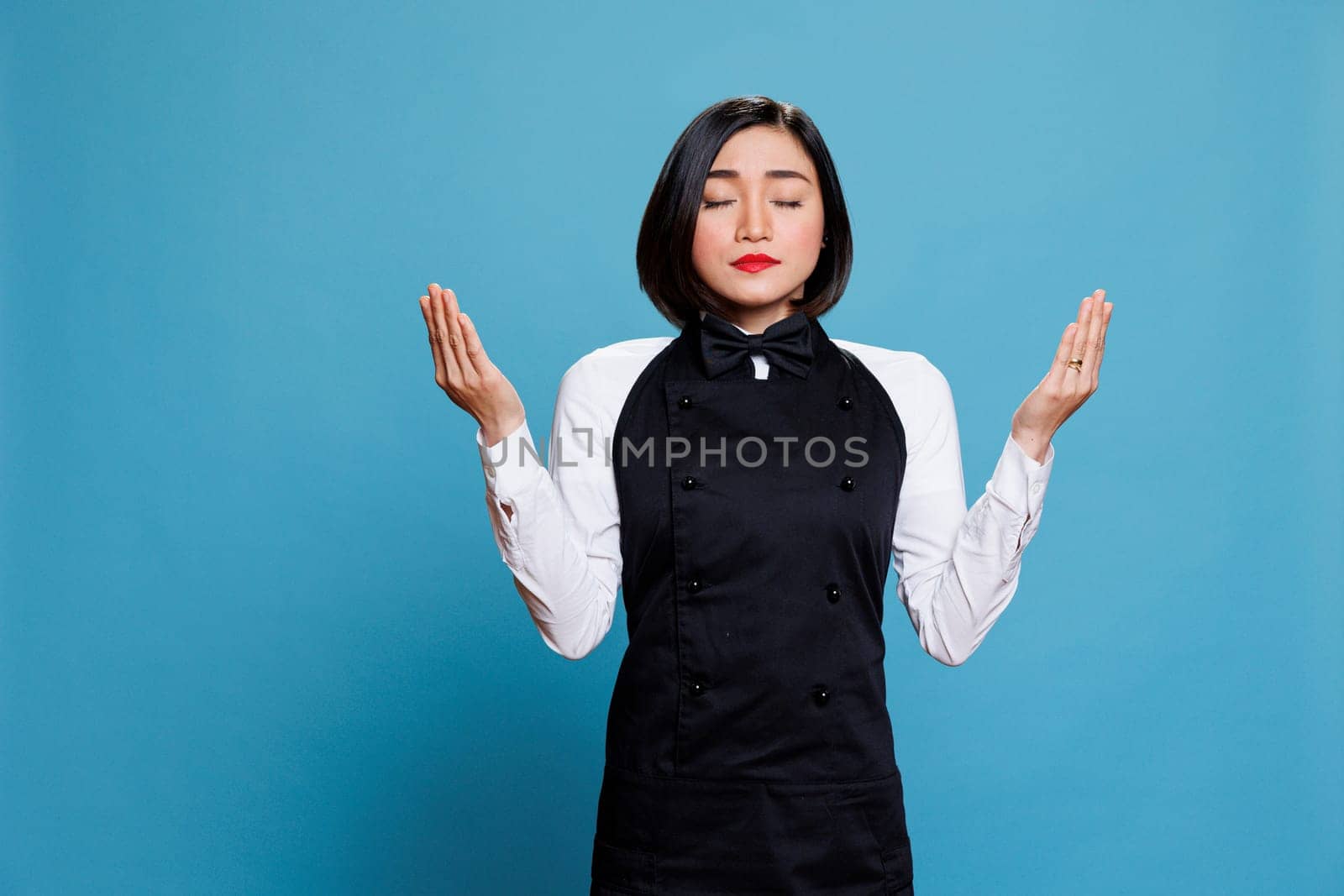 Young attractive asian waitress with closed eyes practicing breathing meditation. Restaurant woman receptionist wearing professional uniform standing with open arms and praying