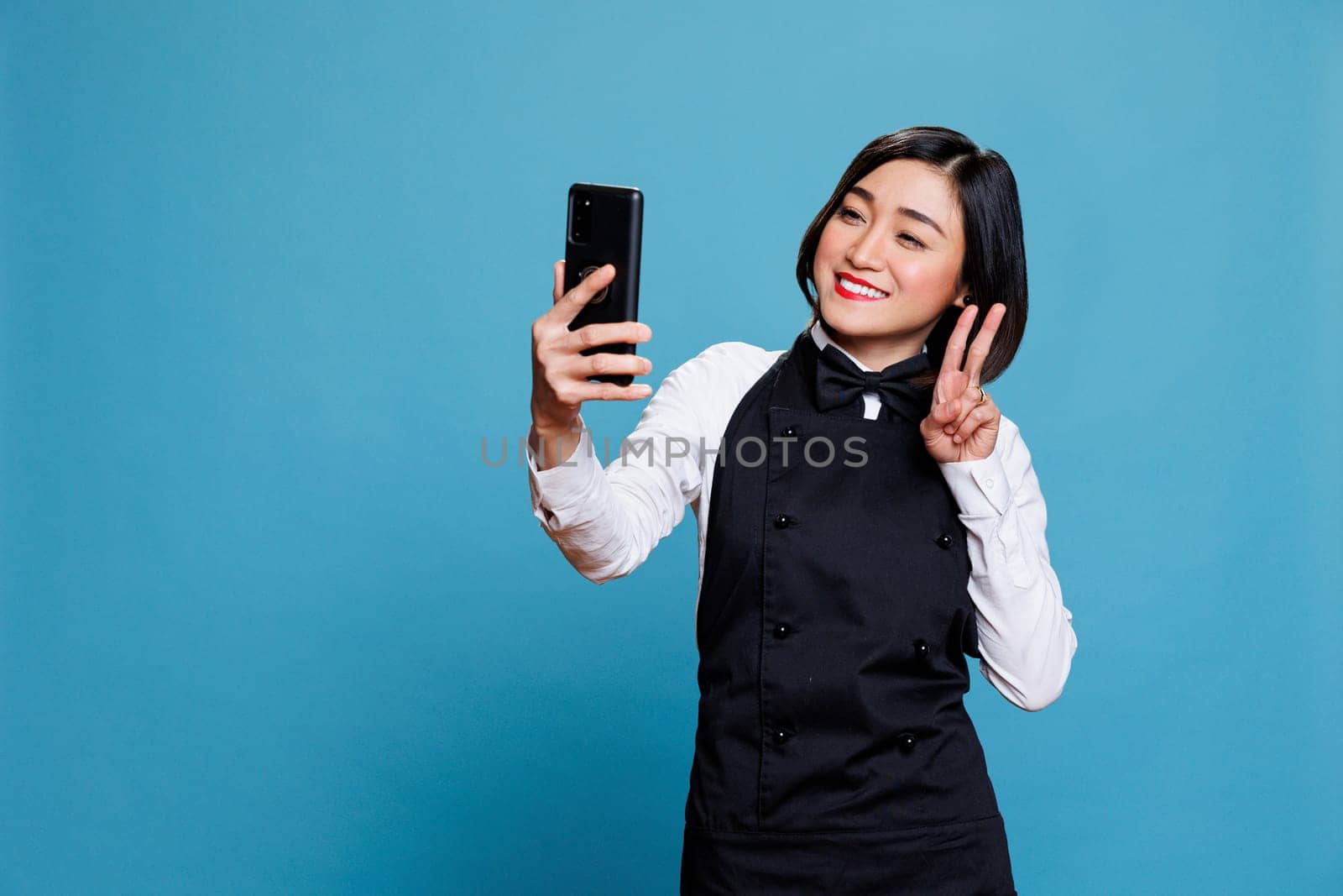 Smiling asian waitress holding smartphone, looking at front camera and showing peace symbol with fingers. Cheerful woman receptionist posing while taking selfie on mobile phone