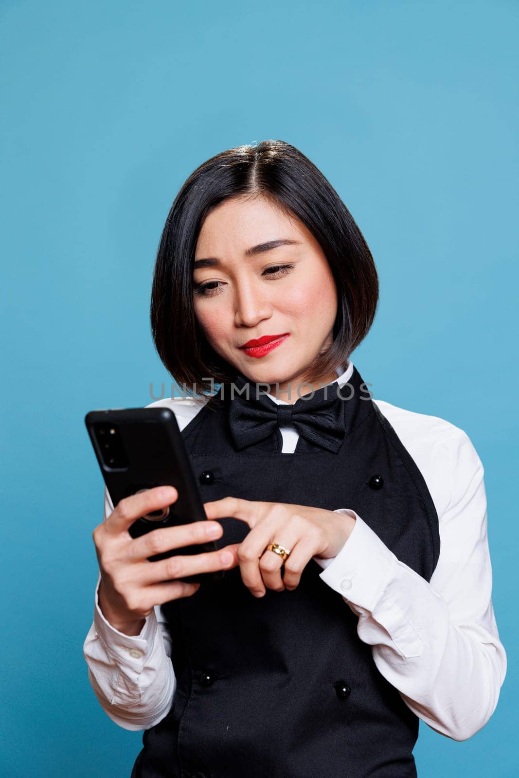 Young smiling asian waitress wearing restaurant uniform tapping on smartphone touchscreen. Attractive woman receptionist answering client message while using mobile phone