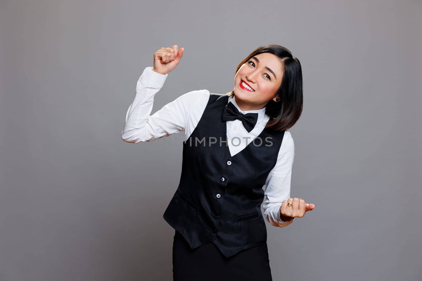 Cafeteria happy waitress wearing black and white professional uniform laughing while dancing. Cheerful young woman receptionist relaxing and having fun, moving arms to music rhythm