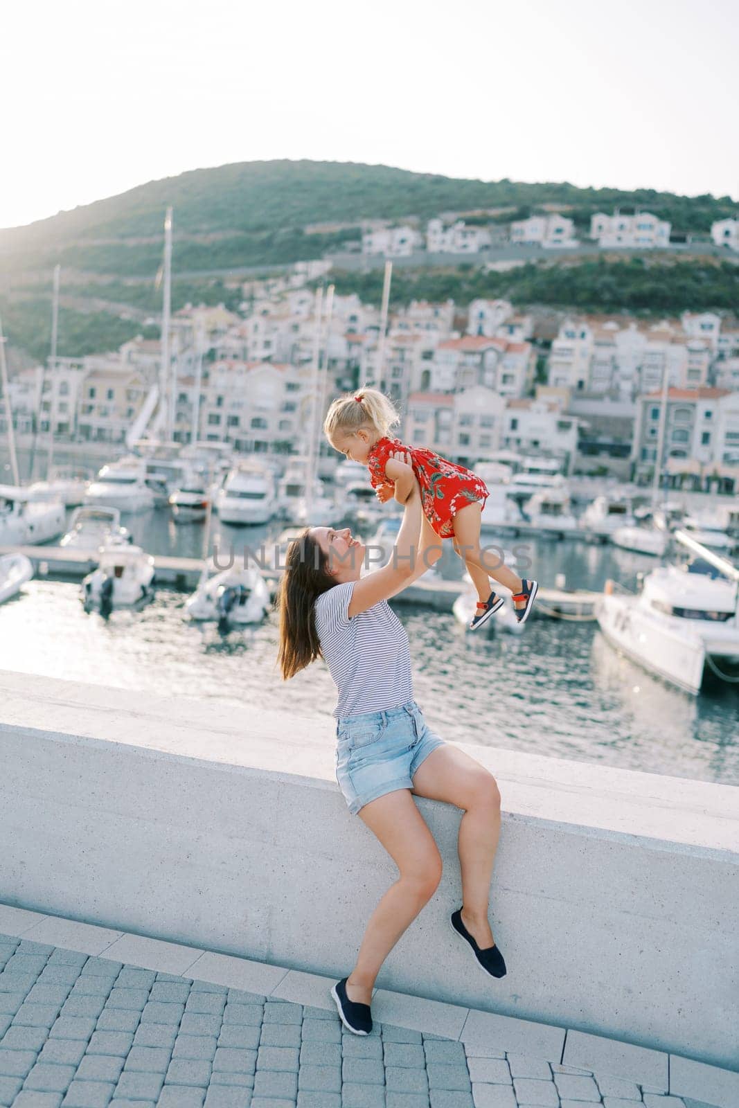 Mom raises a little girl in outstretched arms while sitting on the fence of the boardwalk with moored yachts. High quality photo