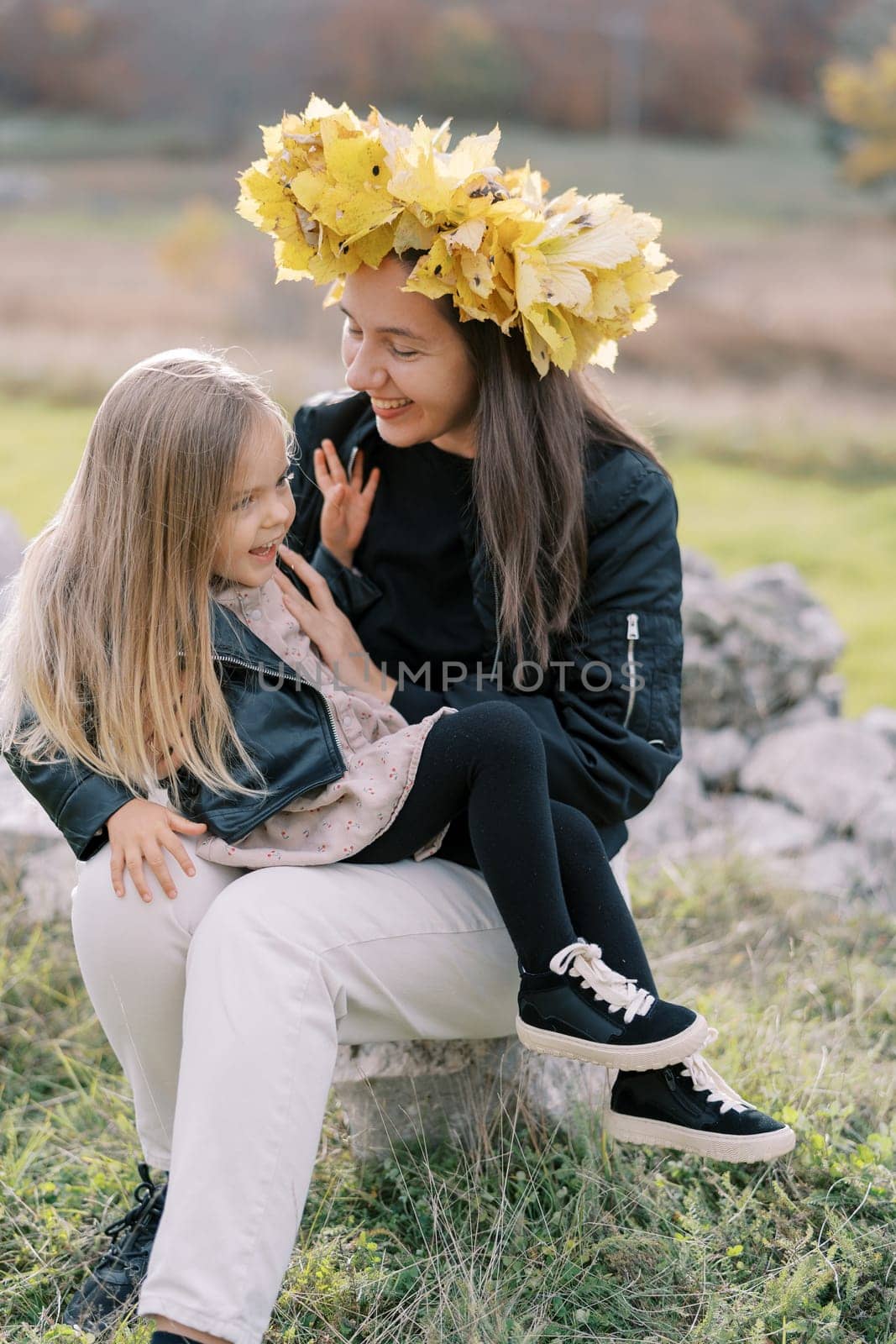 Smiling mother in a wreath of yellow leaves tickles a laughing little girl sitting on her lap on the lawn. High quality photo
