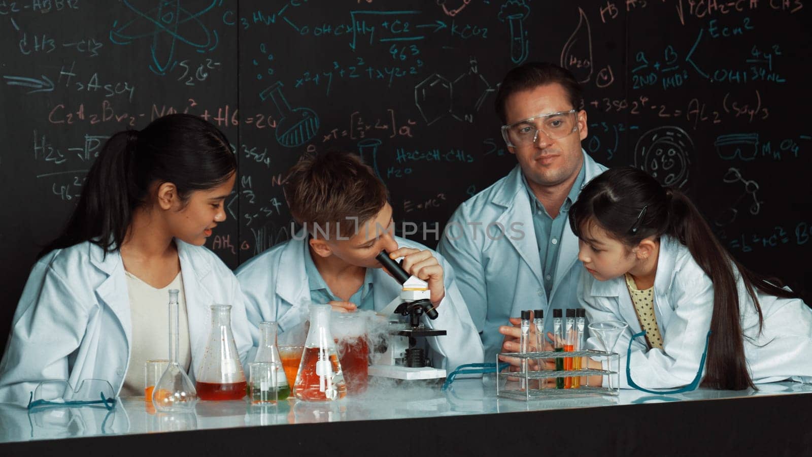 Multicultural student looking under microscope while cute girl inspect sample. Teacher looking after highschool children doing experiment at blackboard with science theory in STEM class. Edification.