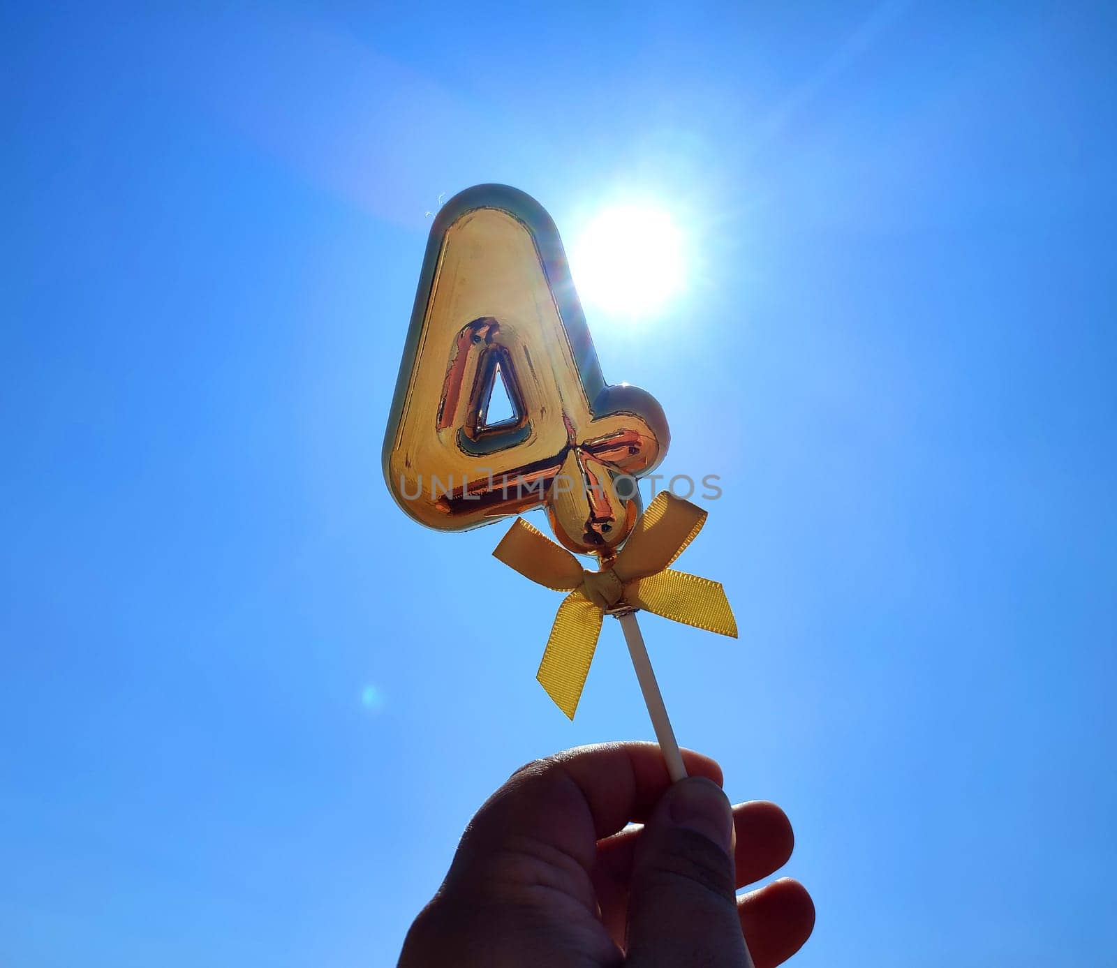 Person holding stick with a shiny golden number 4 four in his hand on backdrop of the waves of blue sky and shining sun on a sunny summer day. Concept holiday birthday party celebrating