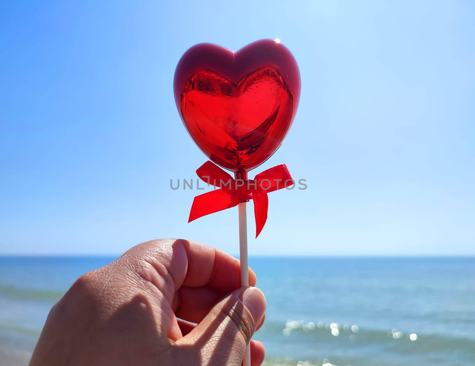 A stick in the shape of a volumetric red heart in woman hand against background of blue sea, sandy beach and blue sky on sunny summer day. Concept love, Valentine Day, falling in love. Vertical