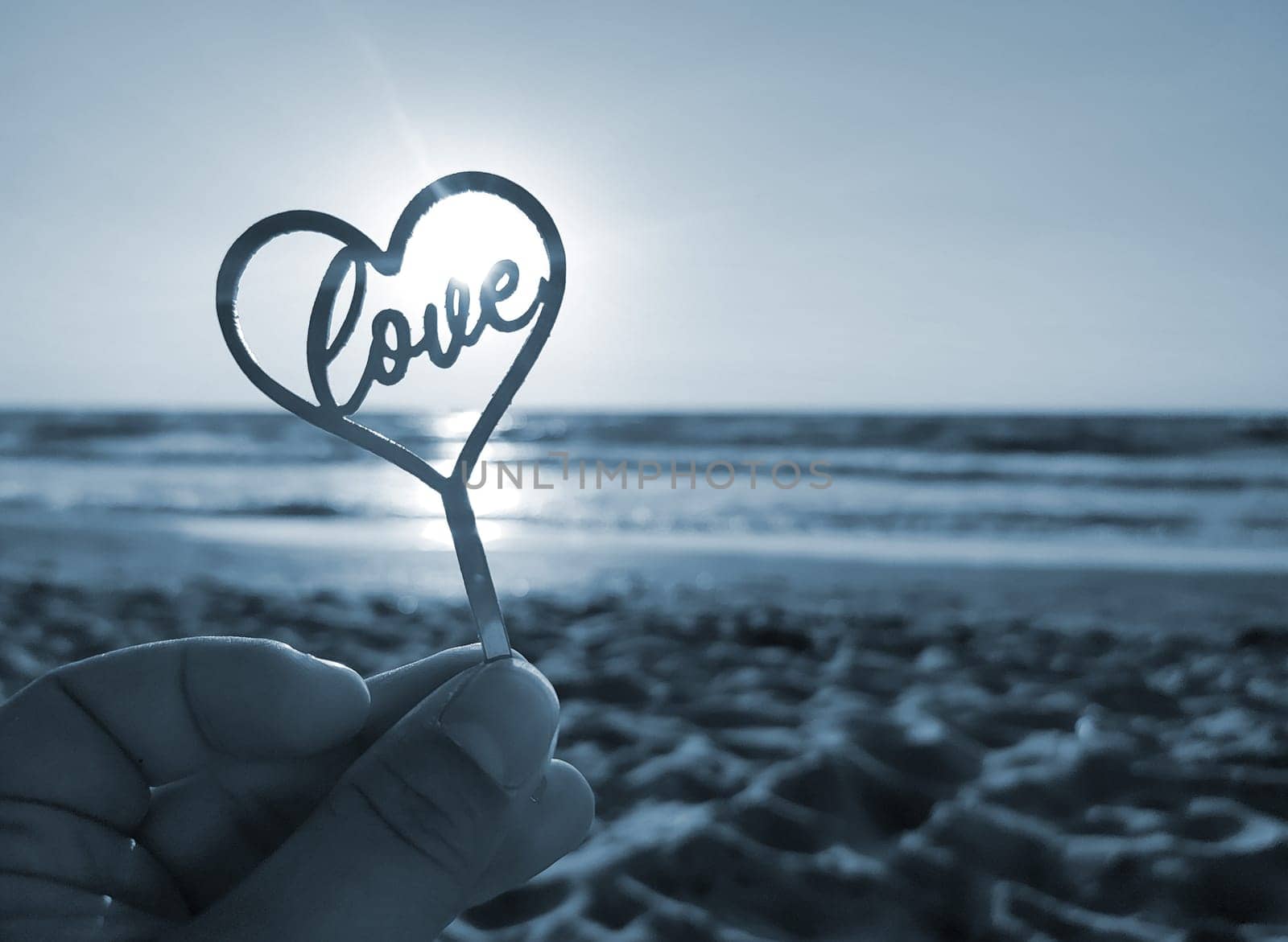 Person holding in fingers hand stick in shape heart and word Love on background sea, waves, blue sky in seashore on sunny summer day close-up. Concept love romance amour St Valentines Day. Blue color