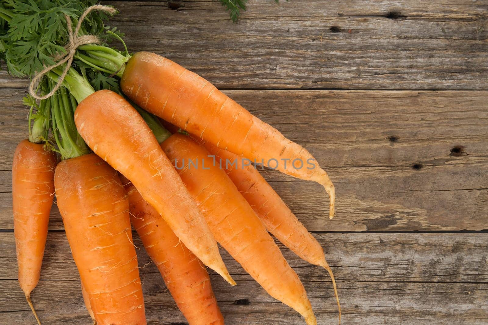 Bunch of fresh, orange carrots with green tops is neatly tied together using twine and positioned on an old, rustic wooden table