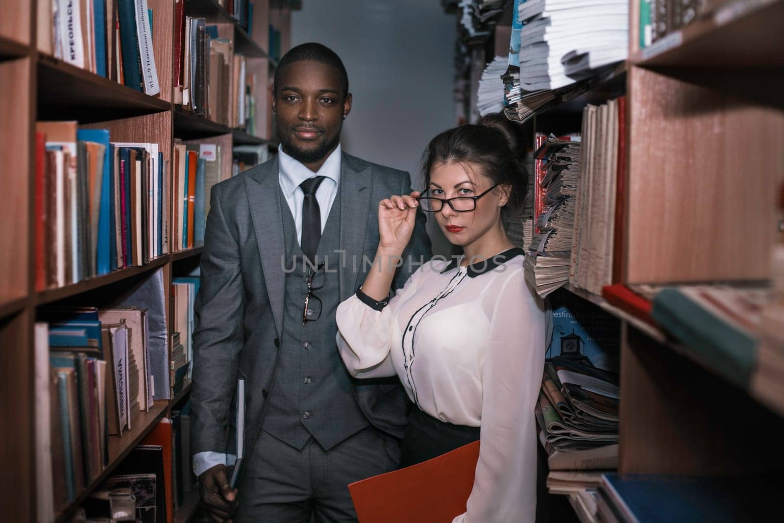 Black man and white girl in the library. The guy is wearing a business suit and the girl is wearing a blouse and eyeglasses.Yaremche, Bukovel, Ukraine - January 10, 2022 by malyshph