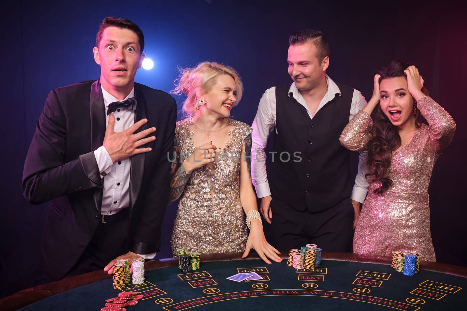 Group of four excited friends are playing poker at casino. Youth are making bets waiting for a big win. They are posing standing at the table against a red and blue backlights on black background. Cards, chips, money, gambling, entertainment concept.