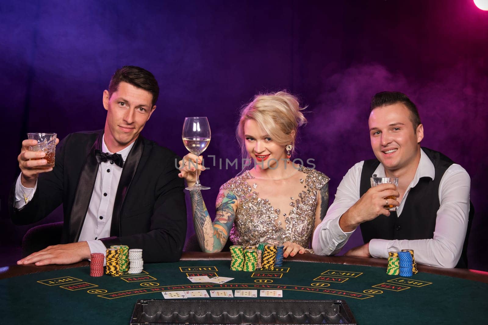 Two stylish men and cute maiden are playing poker at casino. Youth are making bets waiting for a big win. They are smiling and posing sitting at the table against a red and blue backlights on black smoke background. Cards, chips, money, gambling, entertainment concept.