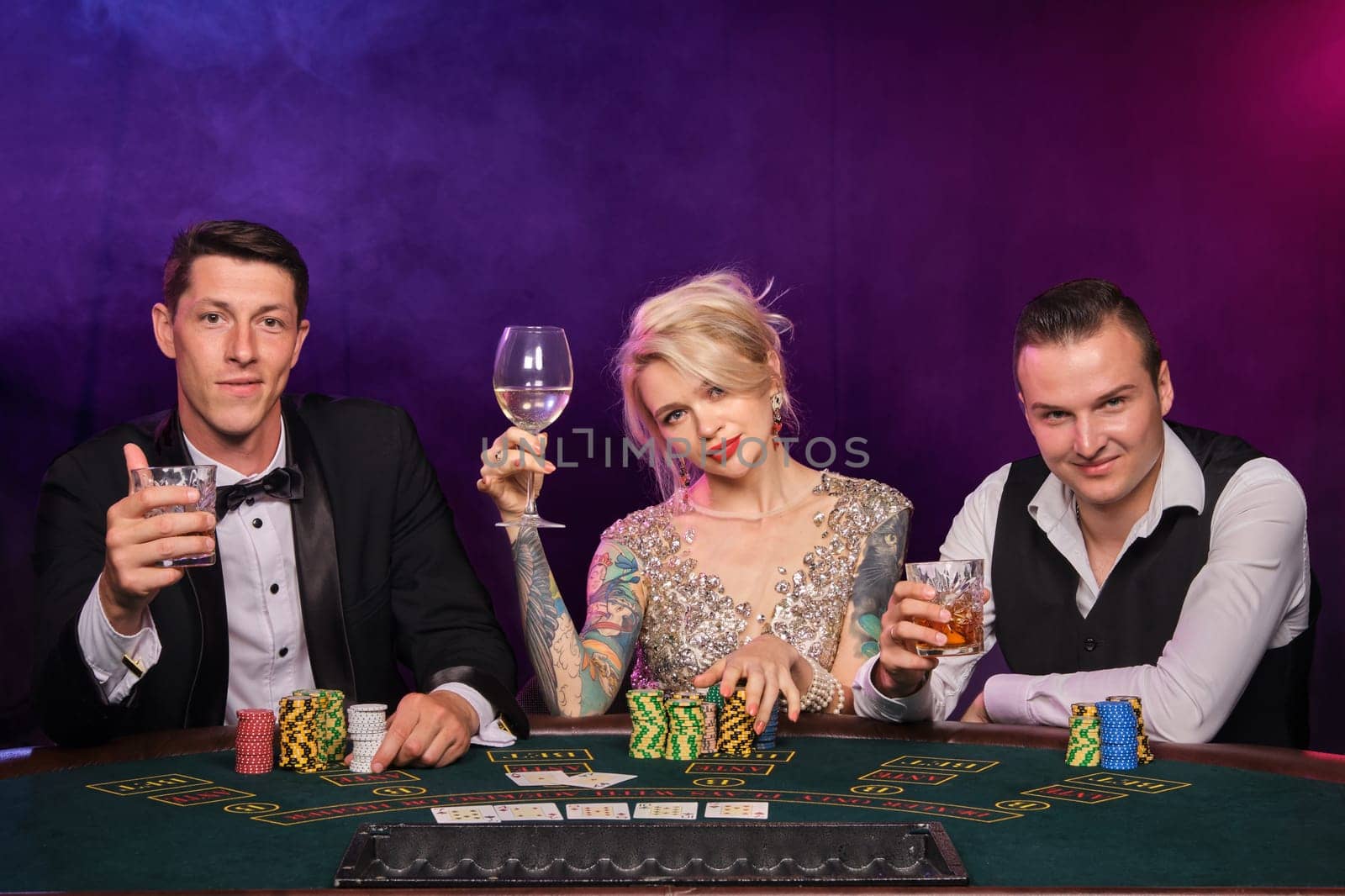 Two stylish fellows and cute lady are playing poker at casino. Youth are making bets waiting for a big win. They are smiling and looking at the camera sitting at the table against a red and blue backlights on black smoke background. Cards, chips, money, gambling, entertainment concept.