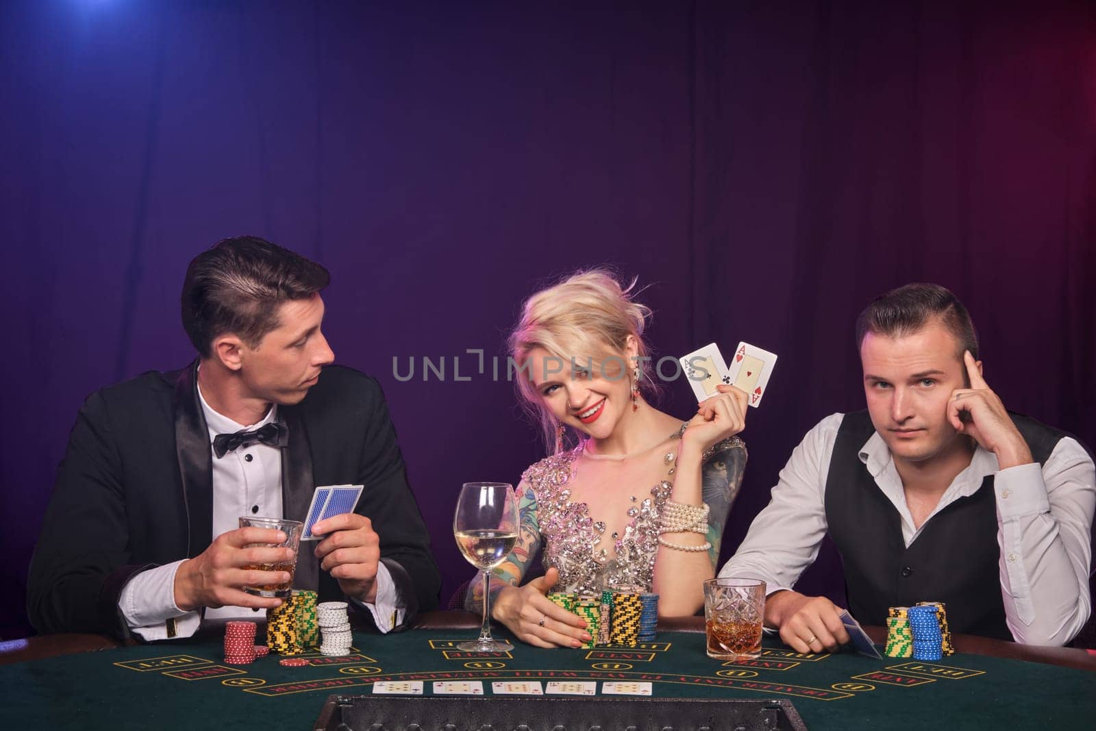 Two stylish men and cute woman are playing poker at casino. Youth are making bets waiting for a big win. They are smiling and posing sitting at the table against a red and blue backlights on black smoke background. Cards, chips, money, gambling, entertainment concept.