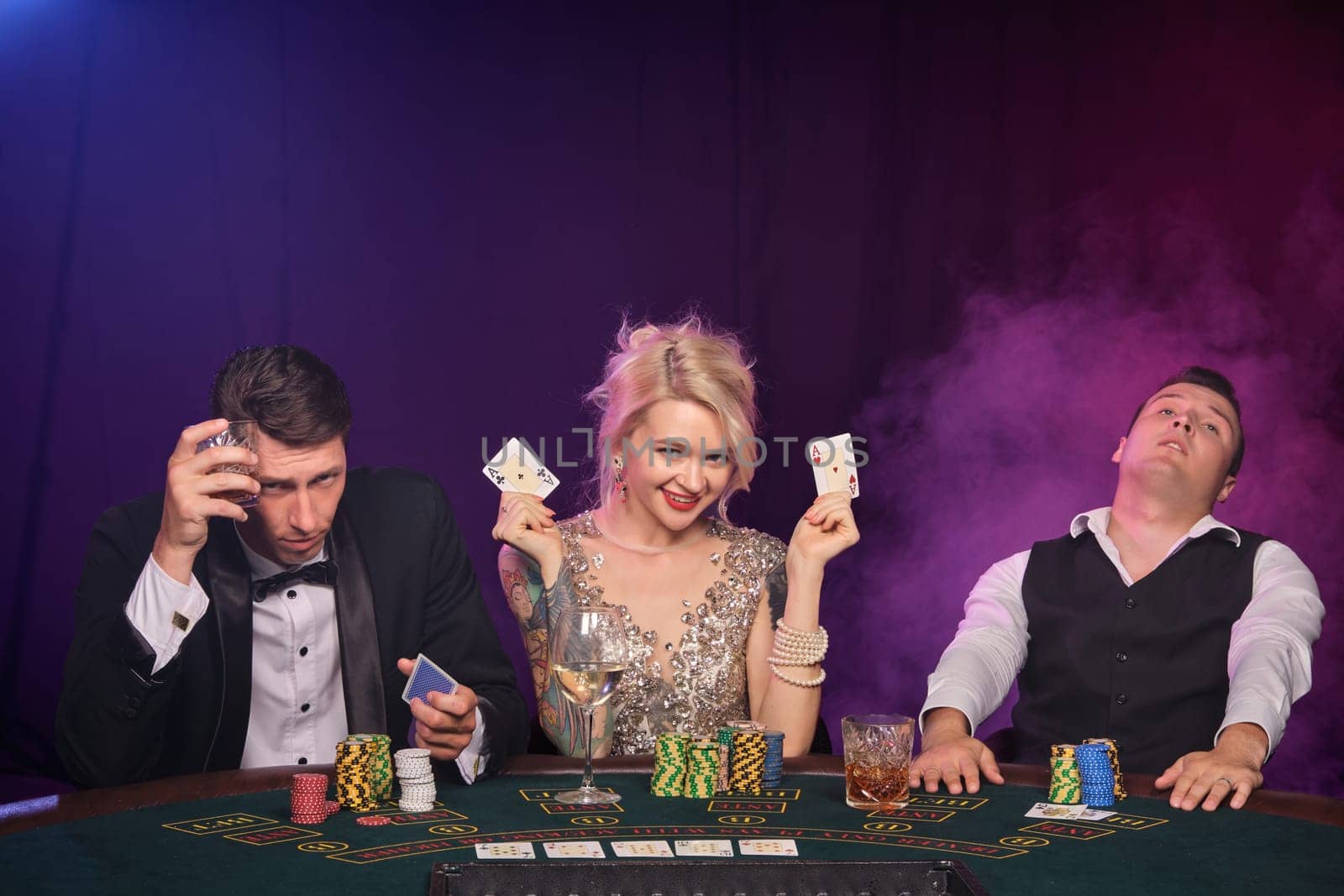 Two stately fellows and pretty lady are playing poker at casino. Youth are making bets waiting for a big win. They posing sitting at the table against a red and blue backlights on black smoke background. Cards, chips, money, gambling, entertainment concept.