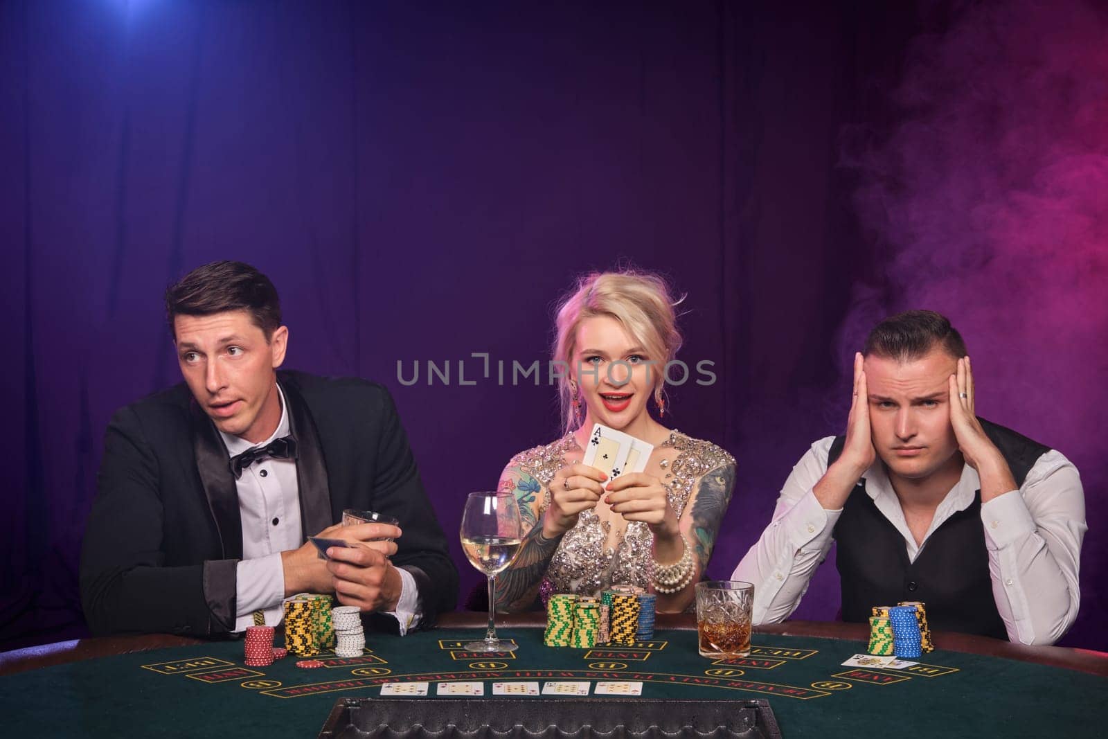Two stately guys and pretty girl are playing poker at casino. Youth are making bets waiting for a big win. They are smiling and posing sitting at the table against a red and blue backlights on black smoke background. Cards, chips, money, gambling, entertainment concept.