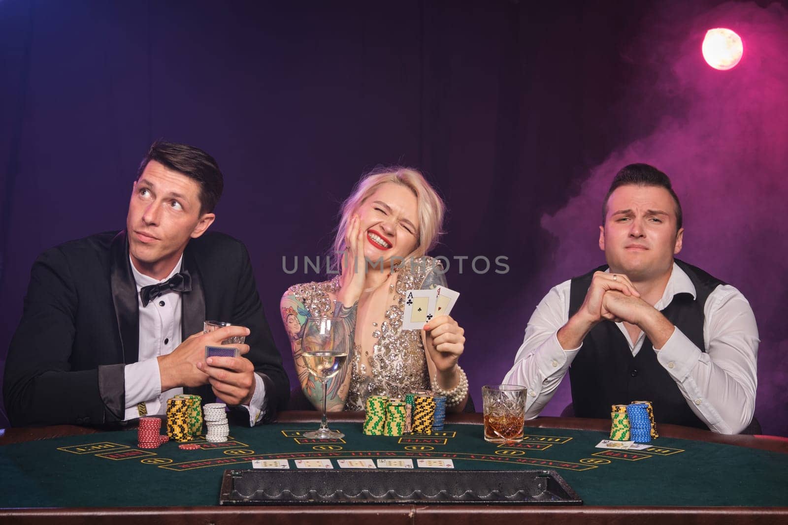 Two stately male and pretty female are playing poker at casino. Youth are making bets waiting for a big win. They are looking upset and posing while sitting at the table against a red and blue backlights on black smoke background. Cards, chips, money, gambling, entertainment concept.