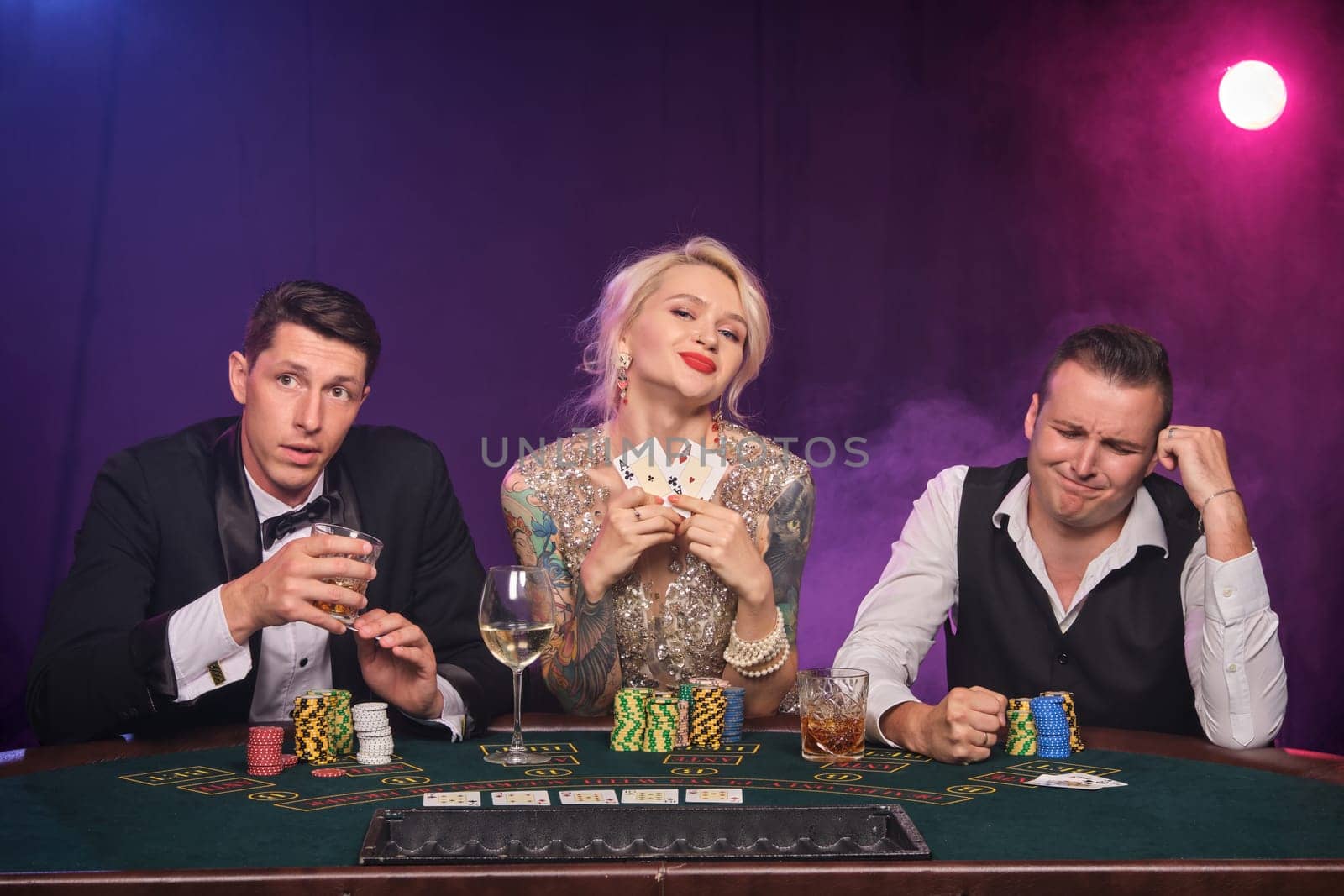 Two rich fellows and elegant lady are playing poker at casino. Youth are making bets waiting for a big win. They are looking unhappy sitting at the table against a red and blue backlights on black smoke background. Cards, chips, money, gambling, entertainment concept.