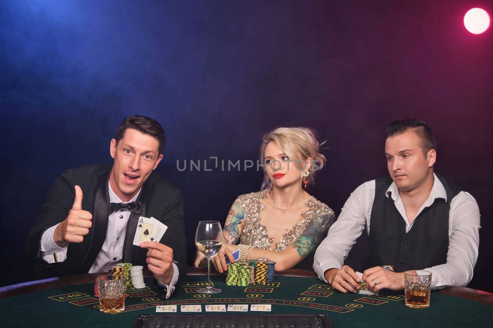 Two wealthy men and charming maiden are playing poker at casino. Youth are making bets waiting for a big win. They are smiling and showing thumb up sitting at the table against a red and blue backlights on black smoke background. Cards, chips, money, gambling, entertainment concept.