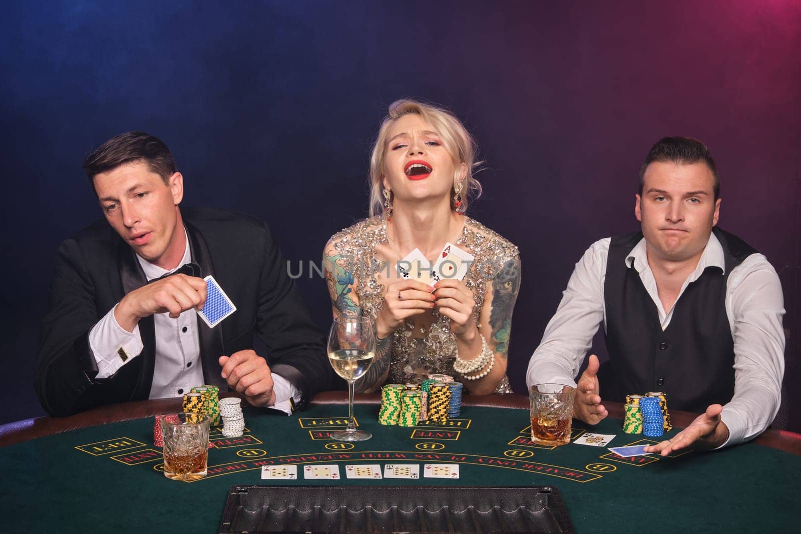 Two rich men and elegant woman are playing poker at casino. Youth are making bets waiting for a big win. They are looking upset sitting at the table against a red and blue backlights on black smoke background. Cards, chips, money, gambling, entertainment concept.