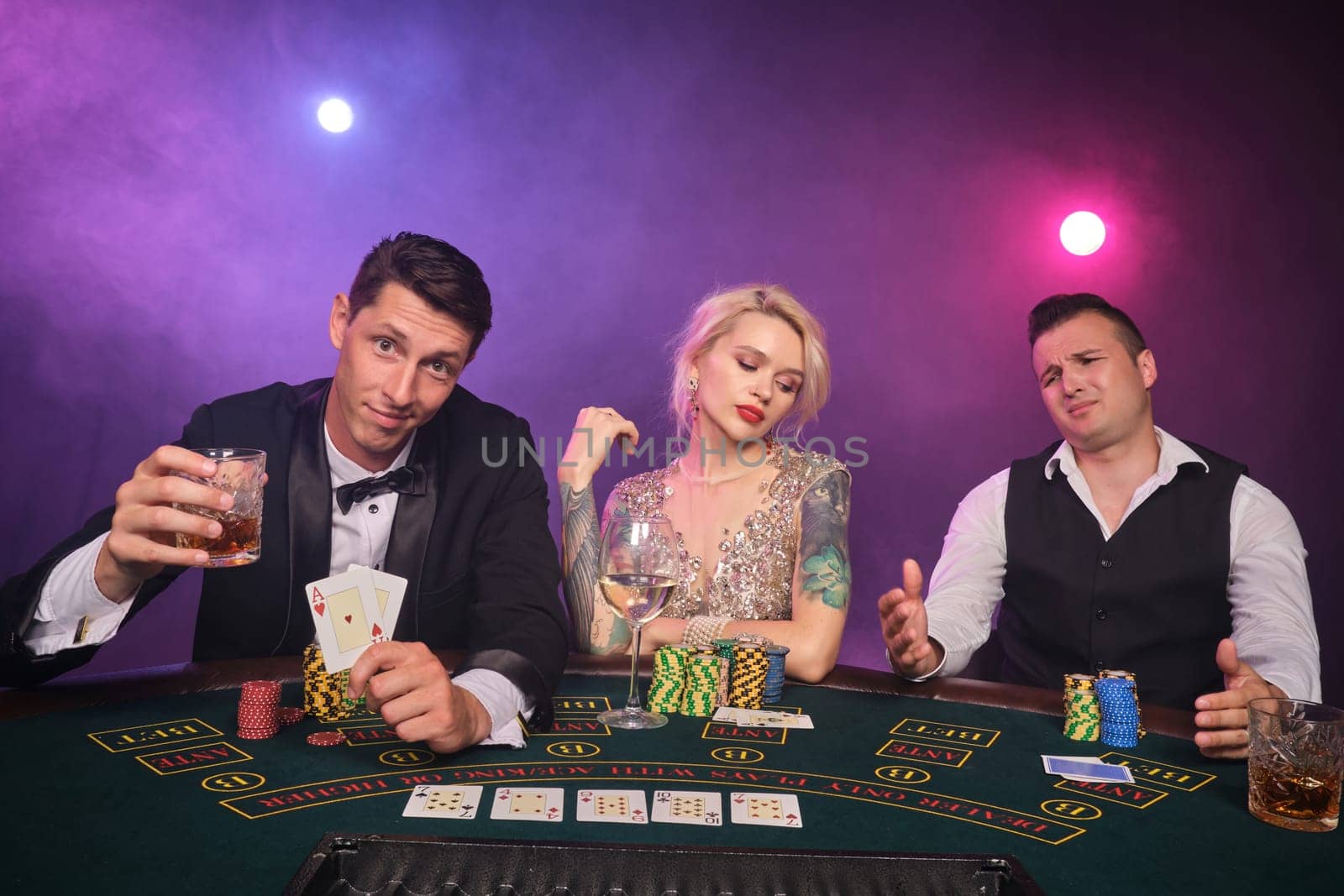 Two wealthy male and charming female are playing poker at casino. Youth are making bets waiting for a big win. They are looking upset and posing sitting at the table against a red and blue backlights on black smoke background. Cards, chips, money, gambling, entertainment concept.