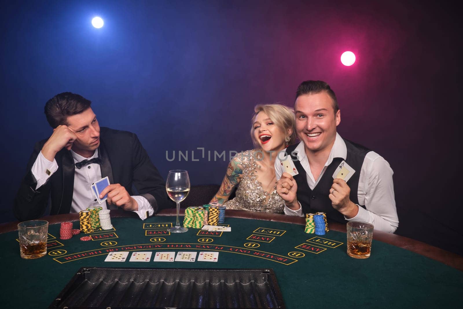 Two athletic men and alluring maiden are playing poker at casino. Youth are making bets waiting for a big win. They are smiling and posing sitting at the table against a red and blue backlights on black smoke background. Cards, chips, money, gambling, entertainment concept.