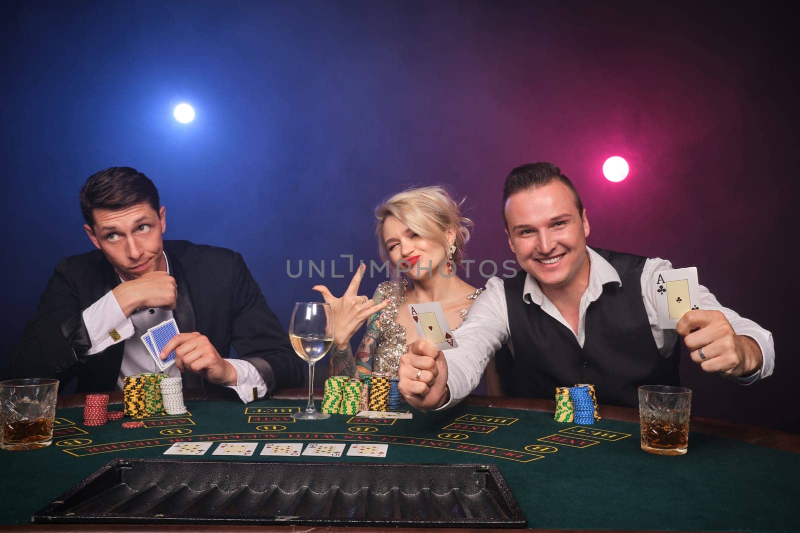 Two athletic guys and alluring girl are playing poker at casino. Youth are making bets waiting for a big win. They are smiling and looking at the camera sitting at the table against a red and blue backlights on black smoke background. Cards, chips, money, gambling, entertainment concept.