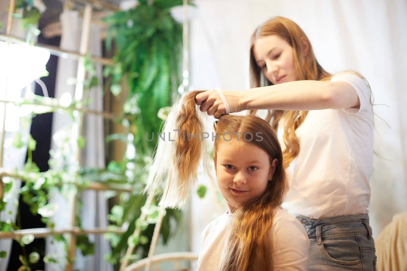 Young loving mom making ponytail to little schoolgirl daughter in living room. Mother helping child girl with hairstyle by keleny