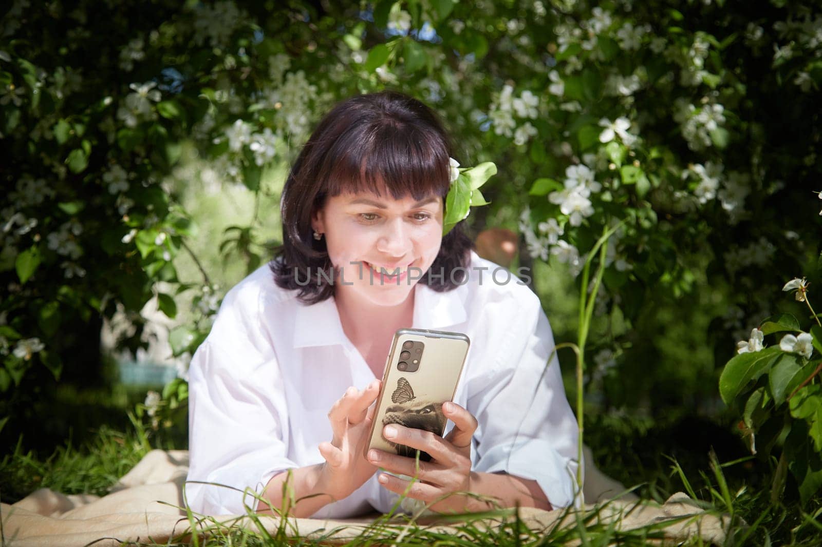 Brunette girl Using Smartphone in Blossoming Orchard in Springtime. Middle aged Woman enjoying phone among spring blossoms of apple or sakura trees