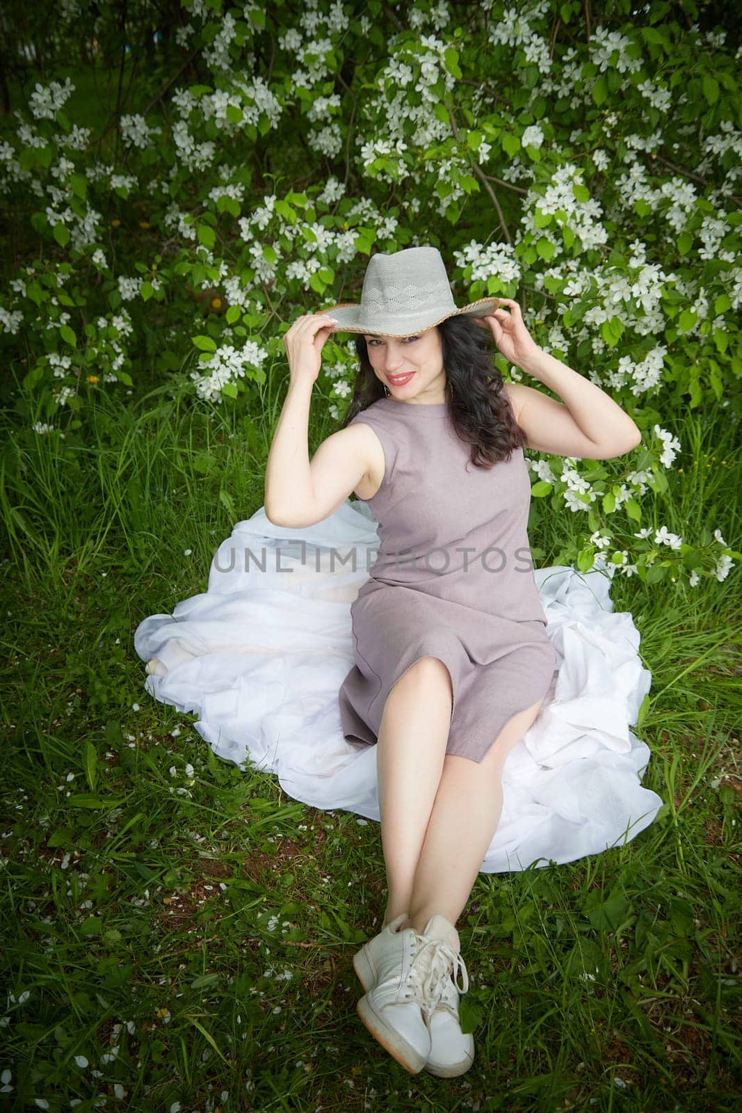 Elegant Woman Posing With Hat in Blossoming Garden. A smiling woman in a dress playfully tipping her hat among spring blossoms. The concept of fashion, self care by keleny