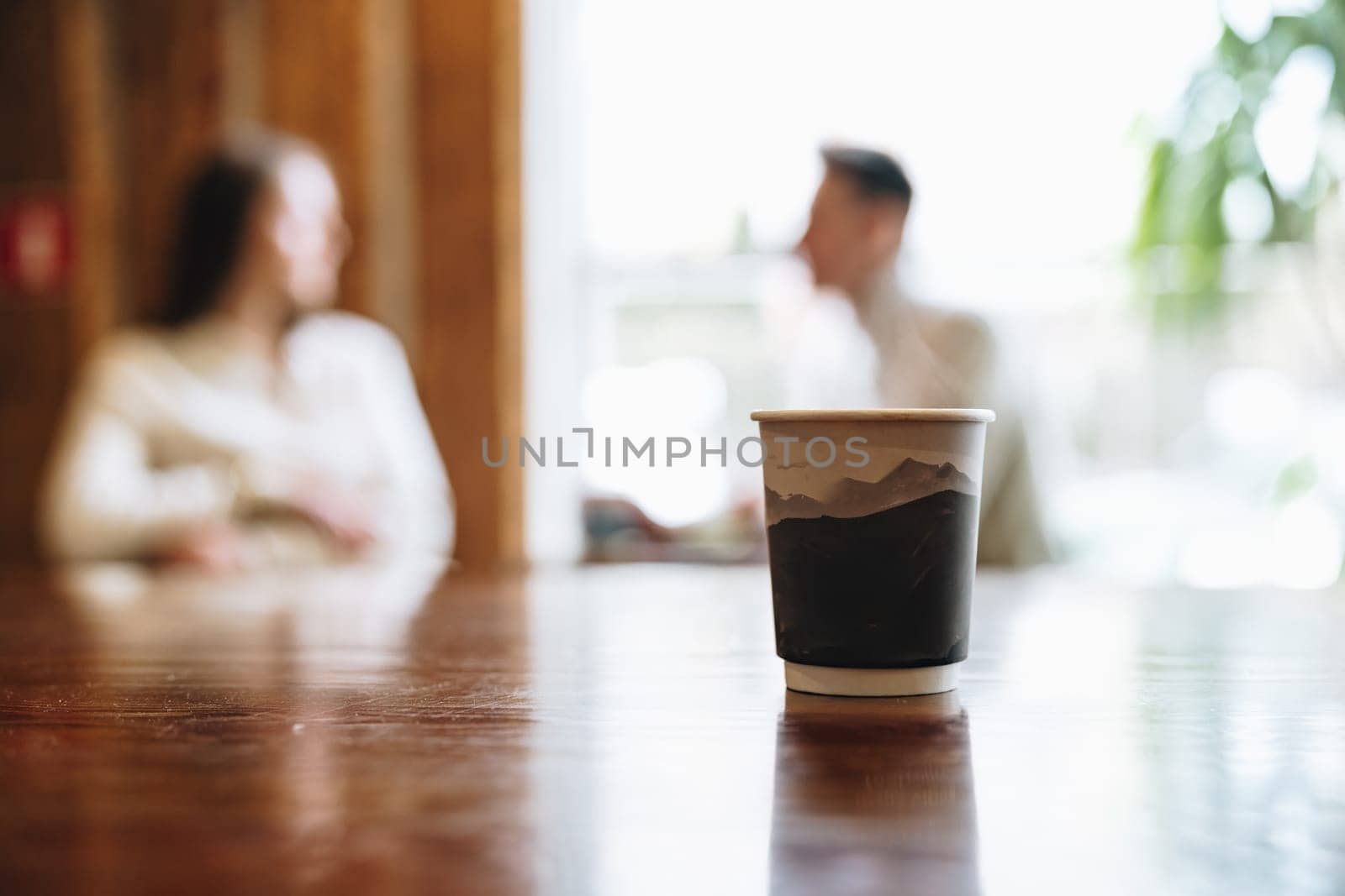 A close-up view of a coffee cup takes the center stage on a wooden table with a blurred background where two people appear to be engaging in a casual conversation in a cozy, indoor setting.