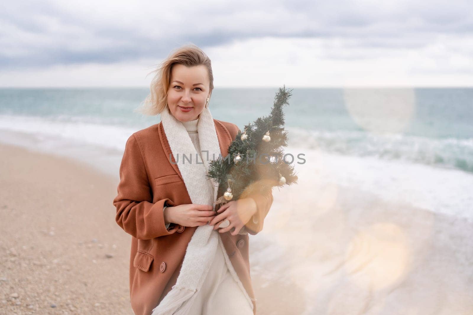 Blond woman Christmas tree sea. Christmas portrait of a happy woman walking along the beach and holding a Christmas tree in her hands. She is wearing a brown coat and a white suit. by Matiunina
