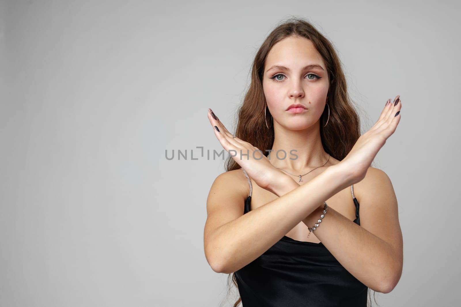 A young woman stands against a muted grey backdrop, her long, wavy hair falling gracefully over her shoulders. She wears a sleek, black sleeveless dress, complementing her fair complexion. With an intense gaze, she crosses her arms to form an X symbol in front of her, suggesting a sign of negation or denial.