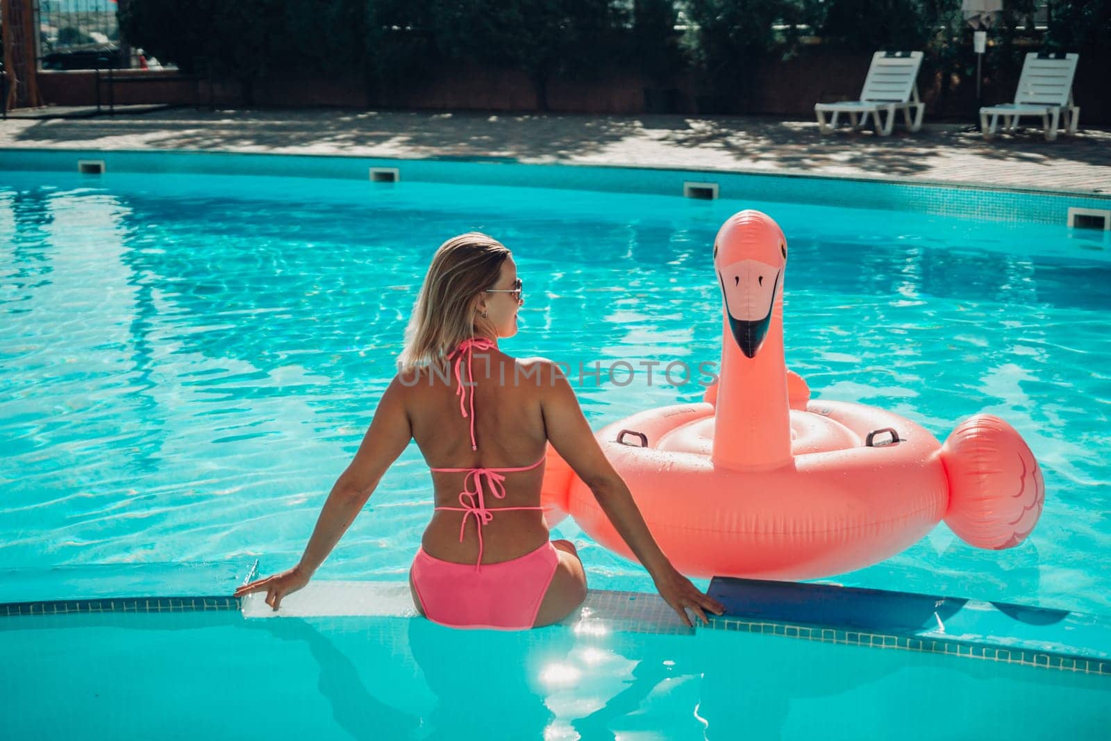 A woman in a pink bikini is sitting on a pink inflatable flamingo in a pool. The scene is playful and fun, with the woman enjoying her time in the water