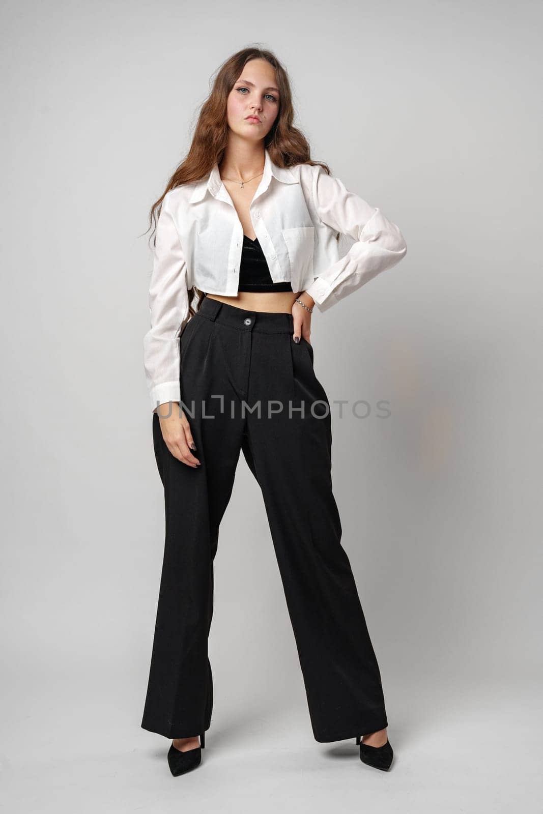 A young woman stands confidently with her hands on her hips, wearing a white blouse partially tucked into high-waisted black trousers, complemented by black heeled shoes. Her direct gaze and natural stance convey self-assurance and fashion sense.