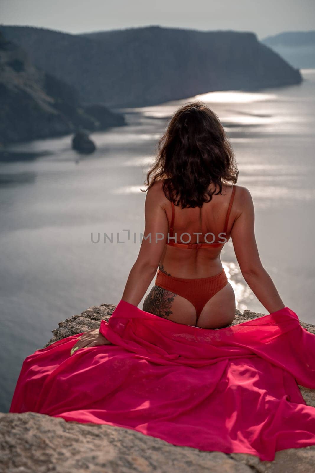 A woman in a red bikini is sitting on a rock overlooking a body of water. The scene is serene and peaceful, with the woman's body language conveying a sense of relaxation and contentment. by Matiunina