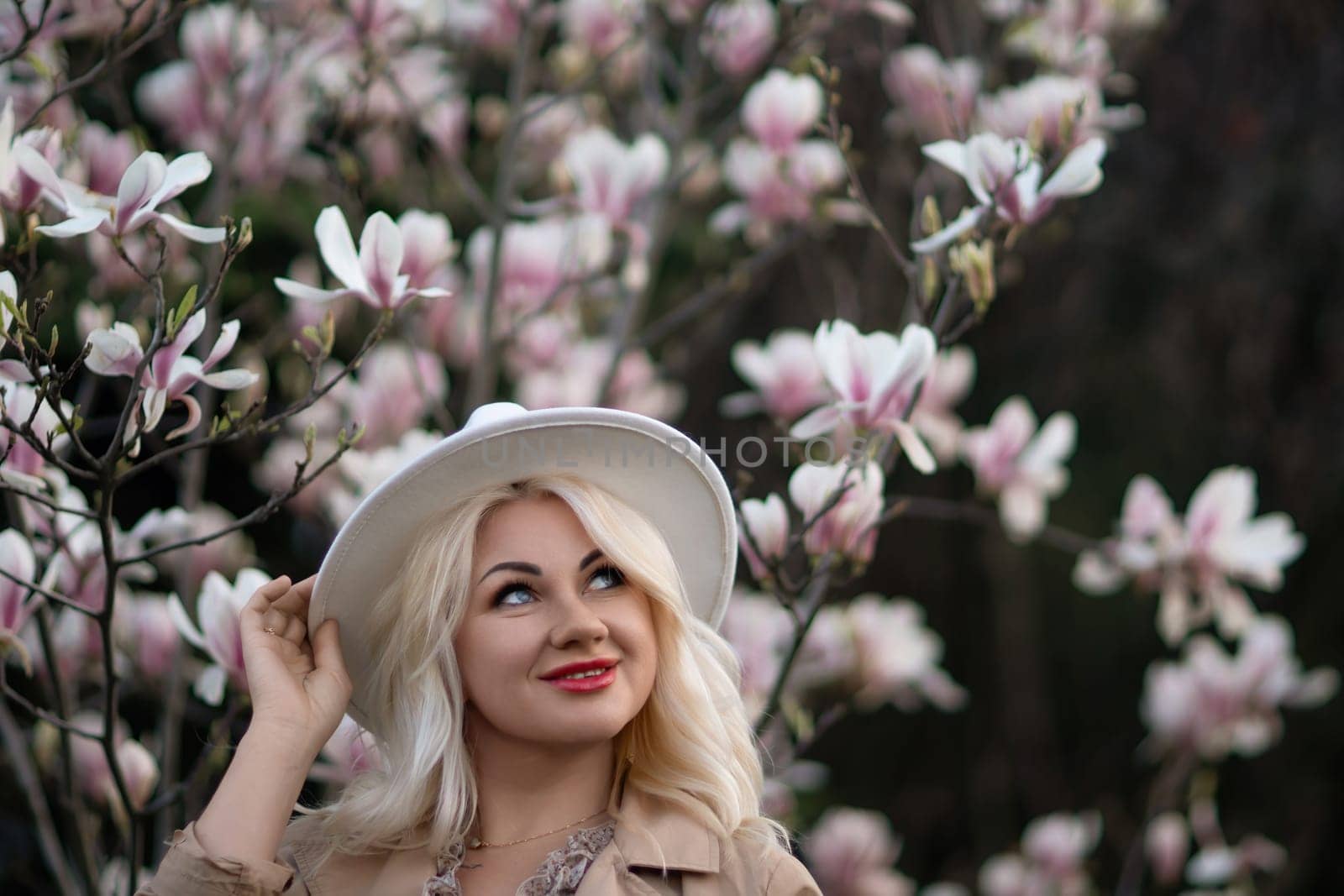 Magnolia flowers woman. A blonde woman wearing a white hat stands in front of a tree with pink flowers. She has a smile on her face and she is enjoying the beautiful scenery. by Matiunina