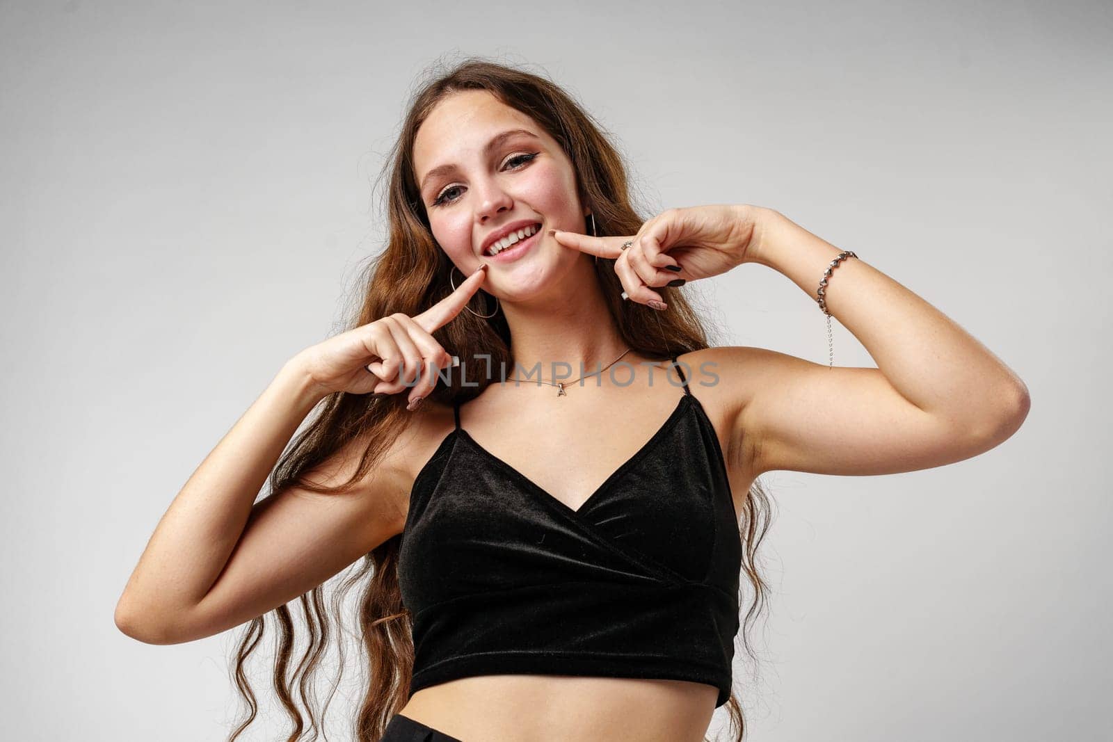 A cheerful young woman with brown hair wearing a black dress is playfully posing against a neutral grey backdrop. She is pointing at her smile, showcasing her happiness, and expressing a positive emotion with sparkling eyes and a beaming expression.