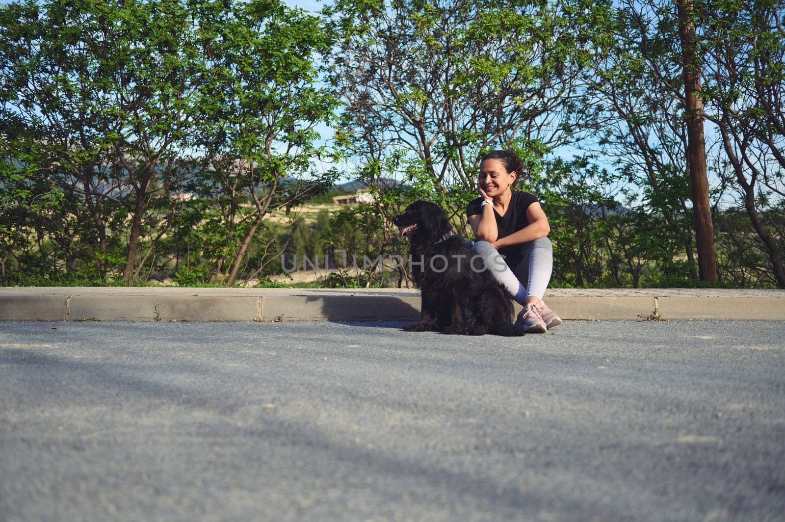 Full size portrait of woman sitting on the parapet, smiling and talking to her dog while taking her pet for a walk on the nature. People and animals concept