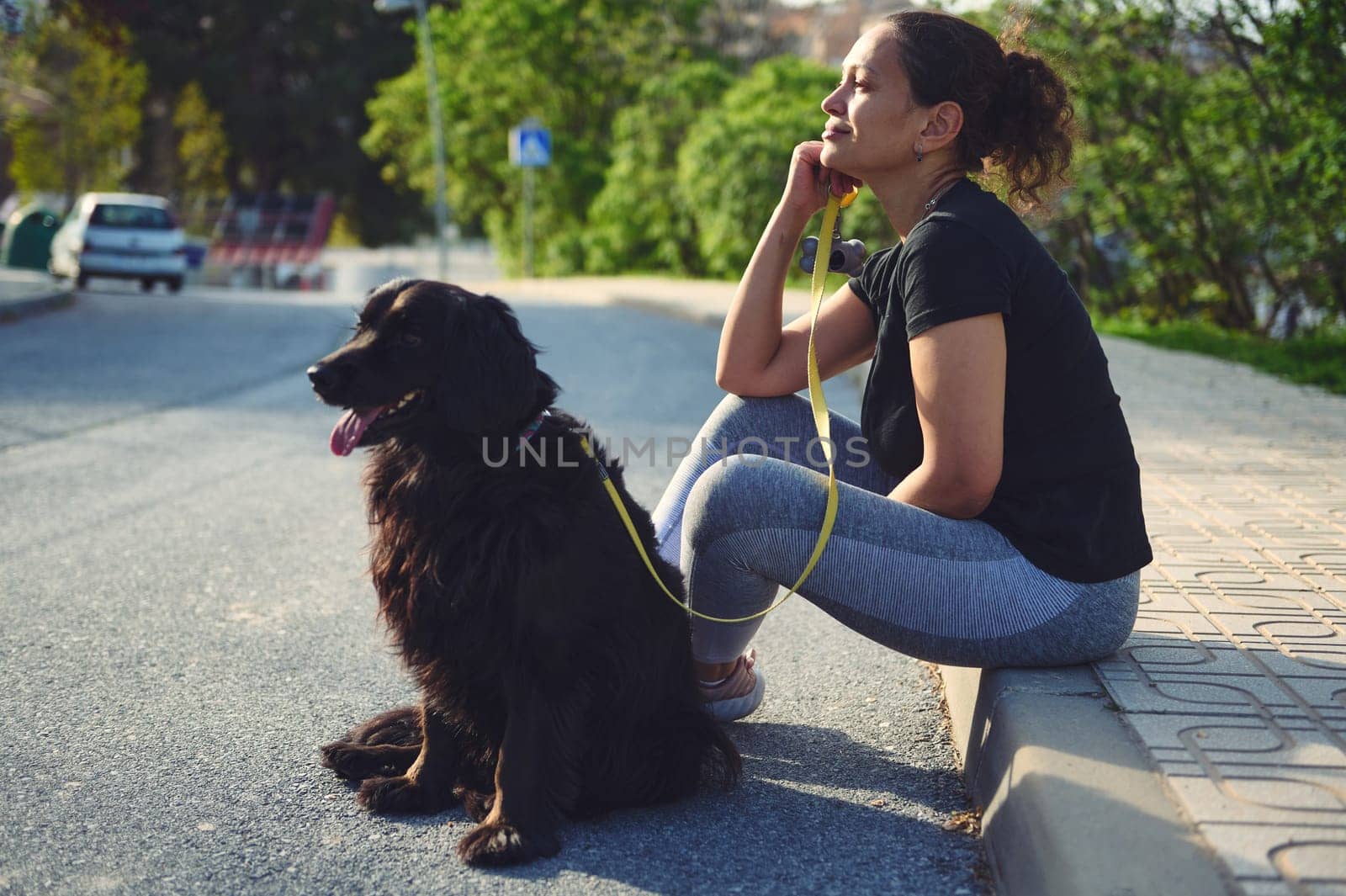 Side portrait of a pensive young adult woman sitting on the parapet, dreamily looking into the distance while taking her pet for a walk on the nature. People and animals concept