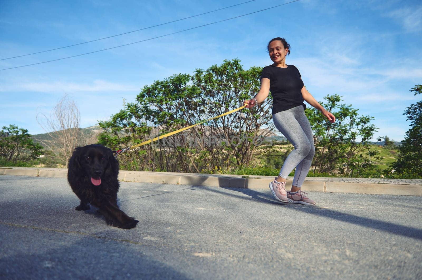 Woman walking her dog on leash in the nature on a beautiful sunny summer day. Pretty female athlete running with her pet in the morning. Playing pets concept by artgf