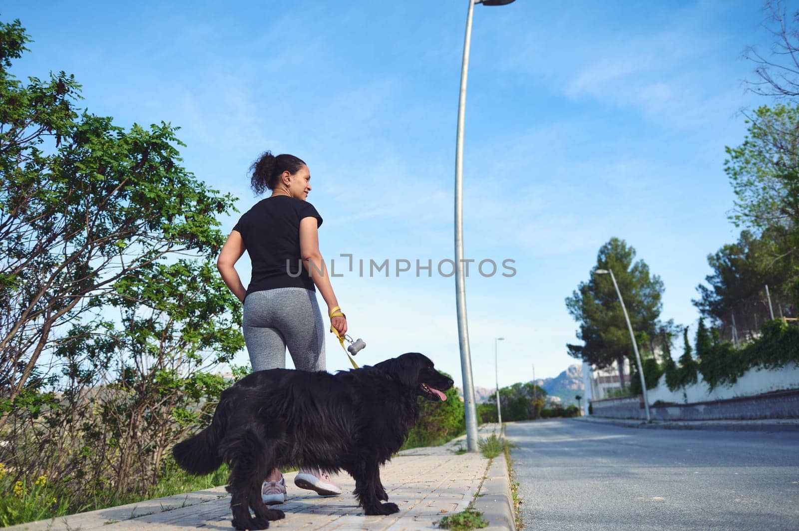 Rear view of a young active woman walking her dog on leash in the nature on a beautiful sunny summer day. Pretty female athlete running with her pet in the morning by artgf