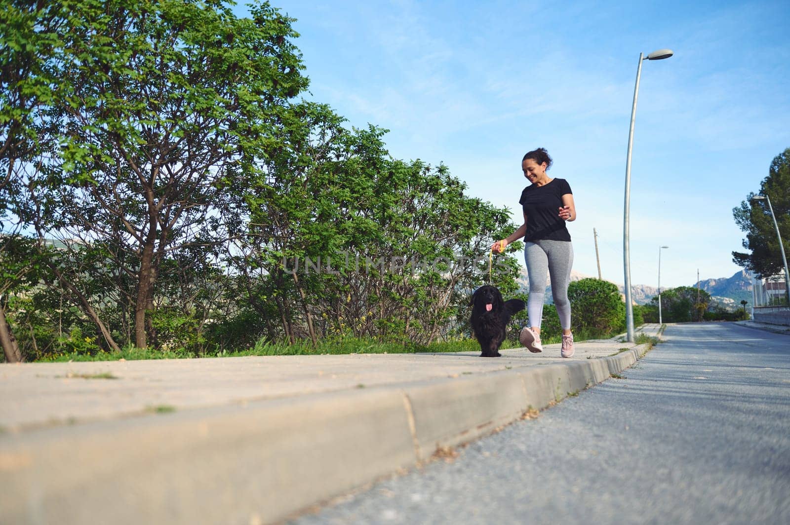 Front view of a young active woman walking her dog on leash in the nature on a beautiful sunny summer day. Pretty female athlete running with her pet in the morning. People and domestic animals