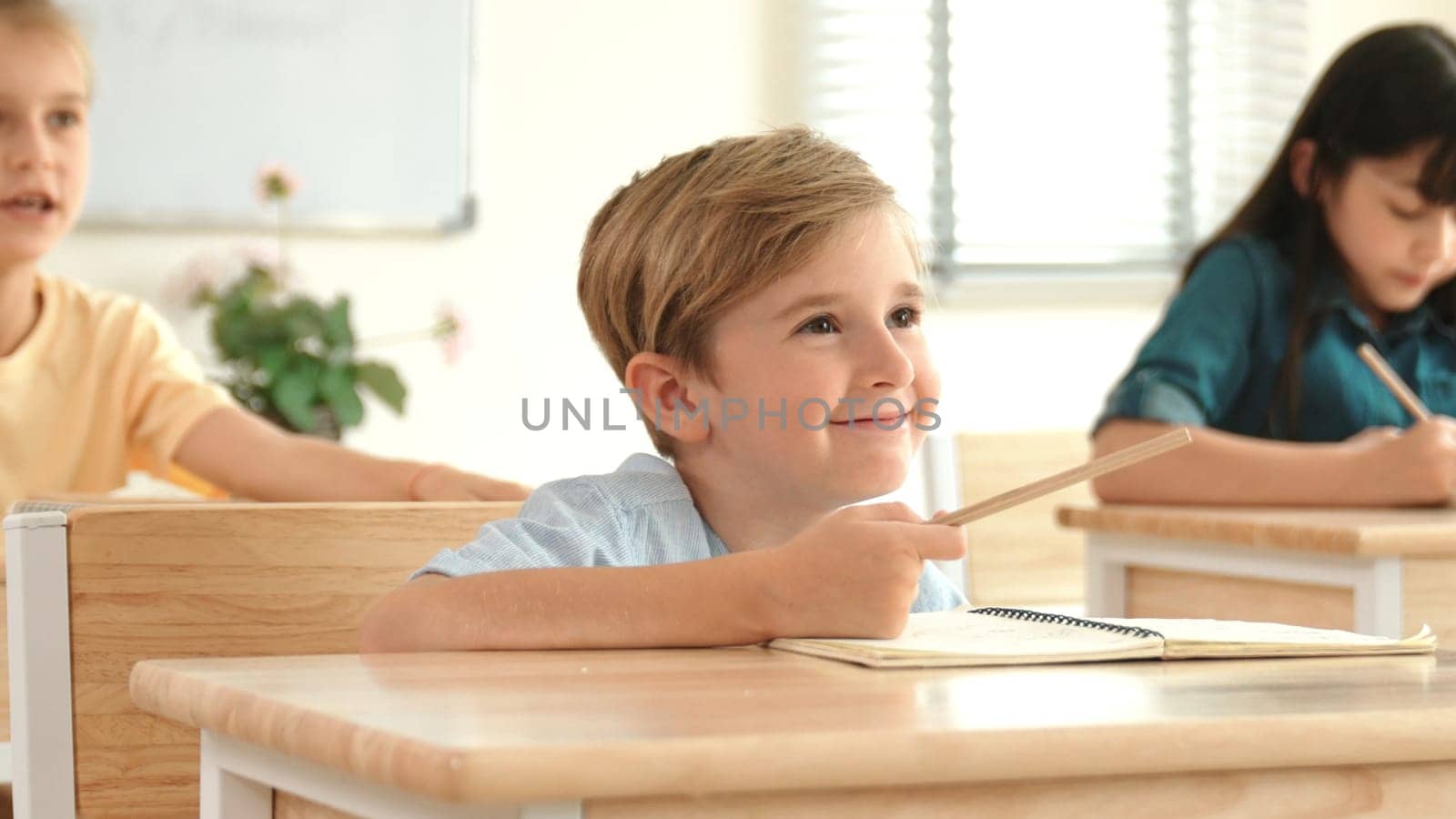 Caucasian boy raising hand for asking and answering teacher surrounded by diverse children studying at classroom. Happy student working together, voting, volunteering, calling instructor. Pedagogy.