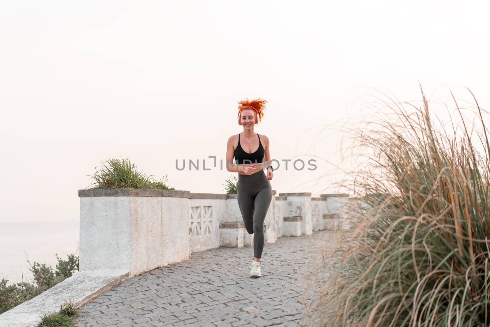 Happy redhead woman running on footpath. Full body of happy fit female runner in sportswear jogging on seaside. Woman jogger listening to music. Health and fitness conepts.
